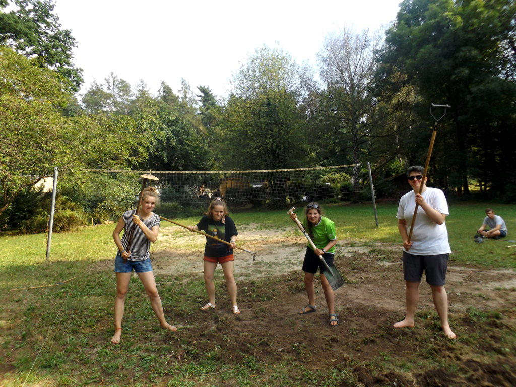 4 Jugendliche halten ihre Arbeitsgeräte hoch, während sie das Volleyballfeld bearbeiten